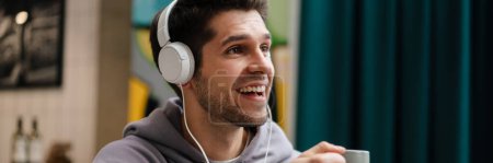 Photo for Smiling young casual man in headphones listening to music, holding mobile phone while sitting in cafe indoors, holding cup - Royalty Free Image