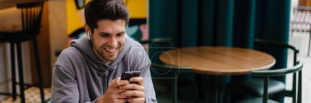 Téléchargez les photos : Joyeux jeune homme brune tenant un téléphone portable tout en étant assis à la table de café à l'intérieur - en image libre de droit