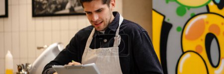 Téléchargez les photos : Jeune serveur vérifiant la qualité des marchandises avec bloc-notes à la cafétéria - en image libre de droit