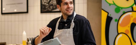 Foto de Sonriente joven camarero comprobando la calidad de las mercancías con bloc de notas en la cafetería - Imagen libre de derechos
