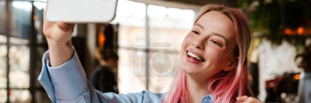 Téléchargez les photos : Jeune belle femme aux cheveux roses prenant selfie photo sur téléphone portable tout en étudiant dans un café - en image libre de droit