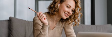 Photo for Young woman in earphones working with laptop while sitting on couch at home - Royalty Free Image