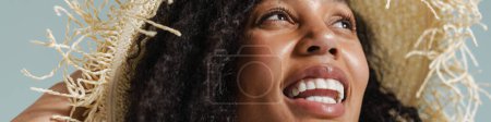 Téléchargez les photos : Femme noire en chapeau de paille souriant et regardant vers le haut isolé sur fond bleu - en image libre de droit
