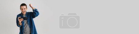 Téléchargez les photos : Jeune homme brune se réjouissant et tenant téléphone cellulaire isolé sur fond blanc - en image libre de droit