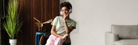 Foto de Young black woman listening music with headphones while sitting in armchair at home - Imagen libre de derechos