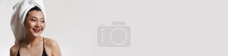 Téléchargez les photos : Brunette asiatique femme portant maillot de bain et serviette souriant à la caméra isolé sur fond blanc - en image libre de droit