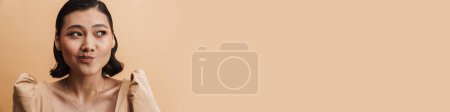 Téléchargez les photos : Jeune femme brune asiatique grimacant et regardant de côté isolé sur fond beige - en image libre de droit