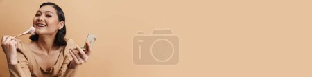 Téléchargez les photos : Jeune femme brune asiatique souriant tout en utilisant une brosse à poudre isolée sur fond beige - en image libre de droit