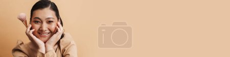 Téléchargez les photos : Jeune femme brune asiatique souriant tout en posant avec brosse à poudre isolée sur fond beige - en image libre de droit
