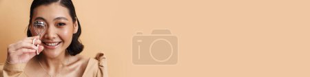 Téléchargez les photos : Jeune asiatique brunette femme souriant tout en utilisant cils curler isolé sur fond beige - en image libre de droit