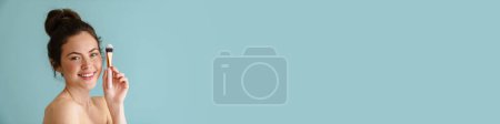 Téléchargez les photos : Femme brune à moitié nue souriant tout en montrant une brosse cosmétique isolée sur fond bleu - en image libre de droit