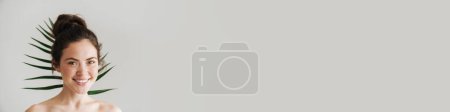 Téléchargez les photos : Femme brune à moitié nue souriant tout en posant avec une feuille verte isolée sur le fond - en image libre de droit