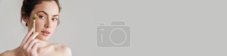 Téléchargez les photos : Femme brune à moitié nue massant le visage avec un rouleau de jade isolé sur le fond - en image libre de droit