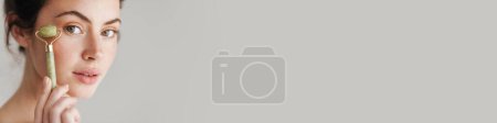 Téléchargez les photos : Femme brune à moitié nue massant le visage avec un rouleau de jade isolé sur le fond - en image libre de droit