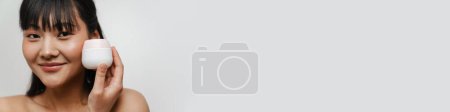 Téléchargez les photos : Jeune asiatique torse nu femme souriant tout en posant avec visage crème isolé sur fond blanc - en image libre de droit