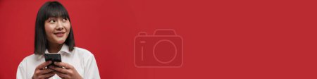 Téléchargez les photos : Jeune femme asiatique portant chemise souriant tout en utilisant un téléphone cellulaire isolé sur fond rouge - en image libre de droit