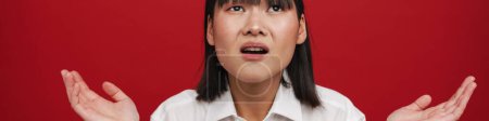 Téléchargez les photos : Jeune femme asiatique portant chemise fronçant les sourcils tout en tenant copyspace isolé sur fond rouge - en image libre de droit