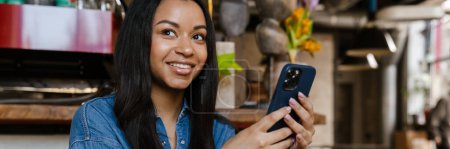 Téléchargez les photos : Femme souriante noire utilisant un téléphone portable alors qu'elle était assise dans un café - en image libre de droit