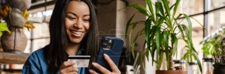 Photo for Black smiling woman using cellphone and credit card while sitting in cafe - Royalty Free Image