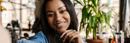 Foto de Negro joven mujer sonriendo y tomando foto selfie mientras está sentado en la cafetería - Imagen libre de derechos