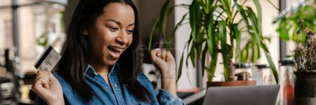 Téléchargez les photos : Noir excité femme en utilisant un ordinateur portable et tenant la carte de crédit tout en étant assis dans le café - en image libre de droit