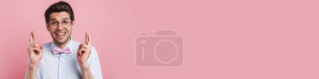 Téléchargez les photos : Portrait d'un jeune homme geek brune souriant portant chemise et noeud papillon debout sur fond mural rose tenant les doigts croisés pour la bonne chance - en image libre de droit