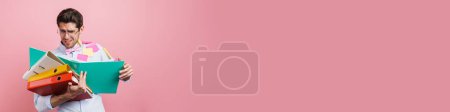 Téléchargez les photos : Stressé homme d'âge moyen employé tenant pile de dossiers avec des documents debout sur fond de mur rose - en image libre de droit