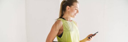 Photo for Young smiling white woman in sportswear carrying fitness mat holding mobile phone standing indoors - Royalty Free Image