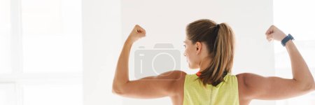 Téléchargez les photos : Femme de fitness souriante montrant ses biceps à la salle de gym devant une grande fenêtre vue arrière - en image libre de droit