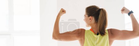 Téléchargez les photos : Femme de fitness souriante montrant ses biceps à la salle de gym devant une grande fenêtre vue arrière - en image libre de droit
