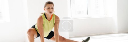 Foto de Estiramiento después de un gran entrenamiento. Joven hermosa mujer joven en ropa deportiva haciendo estiramiento mientras está de pie delante de la ventana - Imagen libre de derechos