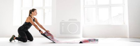 Foto de Joven mujer blanca desenrollando alfombra de fitness antes de practicar en interiores - Imagen libre de derechos