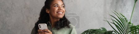 Photo for Black young woman using mobile phone while sitting in armchair indoors - Royalty Free Image