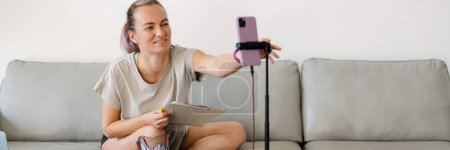 Foto de Una joven blogger blanca discapacitada sonriente con una pierna prostética sentada en un sofá frente a un puesto de cámara haciendo un video en casa - Imagen libre de derechos