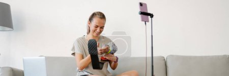 Photo for Smiling young white disabled woman blogger with prostetic leg sitting on a couch in front of a camera stand making a video at home - Royalty Free Image