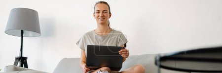 Photo for Smiling young white disabled woman with prostetic leg sitting on a couch with laptop computer at home - Royalty Free Image