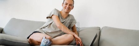 Téléchargez les photos : Sourire jeune femme blanche handicapée avec la jambe prostétique assis sur un canapé avec ordinateur portable à la maison - en image libre de droit
