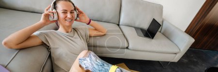 Photo for Top view of a smiling young casual white disabled woman listening to music with headphones sitting on a floor at home leaning on a couch - Royalty Free Image