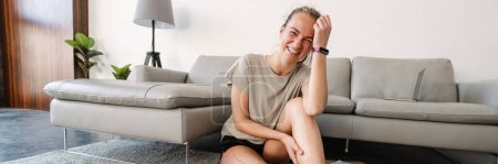 Téléchargez les photos : Jeune femme blanche souriant tout en étant assise avec sa prothèse sur le sol à la maison - en image libre de droit