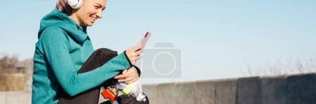 Foto de Joven deportista en auriculares con prótesis sentada en una colchoneta de fitness usando teléfono móvil al aire libre - Imagen libre de derechos