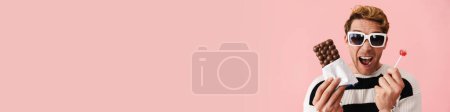 Téléchargez les photos : Heureux mi-âge blonde gay homme en lunettes de soleil debout sur fond rose tenant sucette et chocolat bar gros plan - en image libre de droit