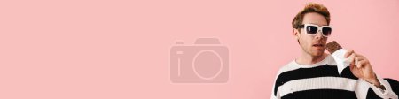 Foto de Feliz mediana edad rubia gay hombre en gafas de sol de pie sobre rosa fondo celebración de chocolate bar - Imagen libre de derechos