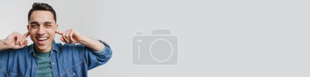 Téléchargez les photos : Jeune homme brune souriant et bouchant ses oreilles isolées sur fond blanc - en image libre de droit