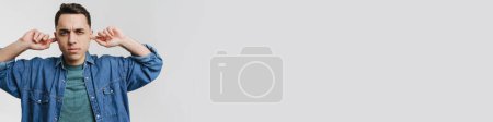 Téléchargez les photos : Jeune homme brune fronçant les sourcils et bouchant ses oreilles isolé sur fond blanc - en image libre de droit