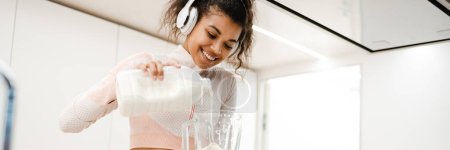 Photo for Black woman in headphones smiling and making smoothie with milk at home - Royalty Free Image
