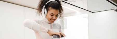 Photo for Black woman in headphones smiling and making smoothie in kitchen at home - Royalty Free Image