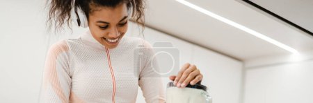 Photo for Black woman in headphones smiling and making smoothie at home - Royalty Free Image