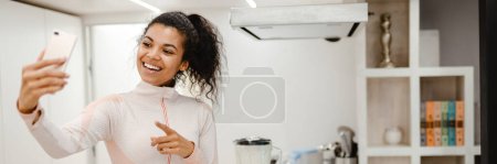 Photo for Black woman taking selfie on mobile phone while cooking in kitchen at home - Royalty Free Image