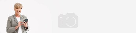 Téléchargez les photos : Européenne mature femme en veste souriant tout en utilisant un téléphone mobile isolé sur fond blanc - en image libre de droit