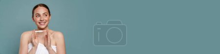 Téléchargez les photos : Femme roux portant une serviette souriante et montrant test de grossesse isolé sur fond bleu - en image libre de droit